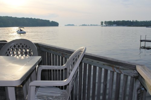 A serene lakeside view with a table and chair on a deck, a boat in the distance, and trees lining the shore.