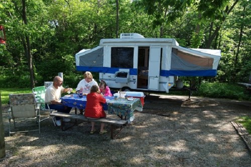 A family enjoys a meal at a picnic table near a pop-up camper in a wooded campsite.