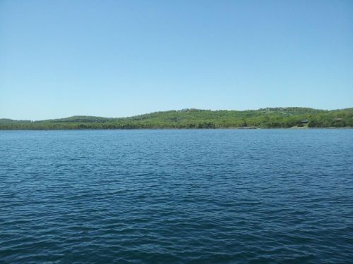 A serene view of a calm blue lake with lush green hills in the background under a clear blue sky.