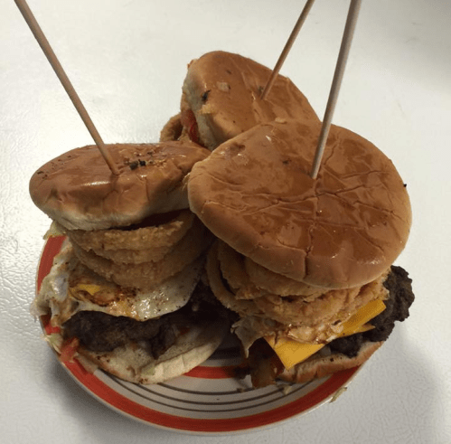 A stack of burgers with onion rings, cheese, and toppings, held together by toothpicks on a plate.