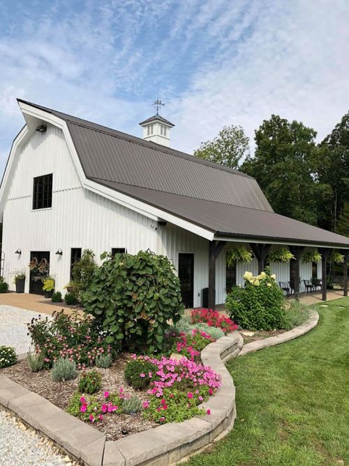 A charming white barn with a metal roof, surrounded by colorful flowers and greenery under a partly cloudy sky.