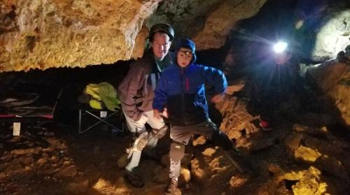 Two people in a cave, one crouching and the other standing, with a flashlight illuminating the rocky surroundings.