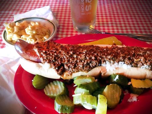 A plate with a chili dog topped with onions, pickles, and a side of macaroni and cheese, with a drink in the background.