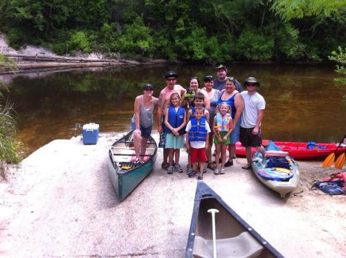 A group of people, including children, poses by a river with canoes and kayaks on a sunny day.