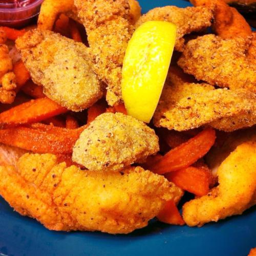 A plate of golden fried fish and shrimp, garnished with a lemon slice and served with sweet potato fries.