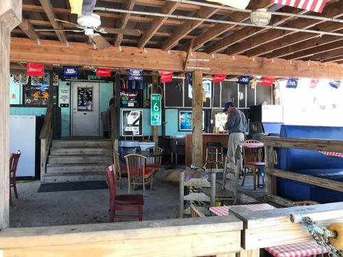 A rustic bar with wooden seating, a TV, and a man cleaning near the entrance, decorated with beer signs and flags.