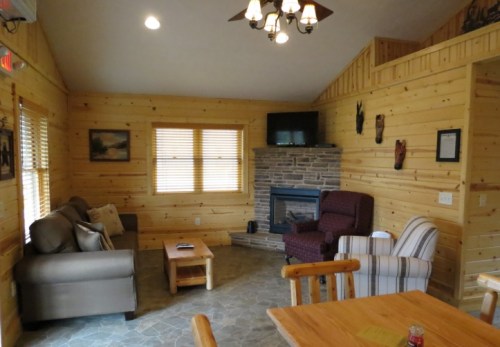 Cozy cabin interior with wooden walls, a stone fireplace, comfortable seating, and a dining table.