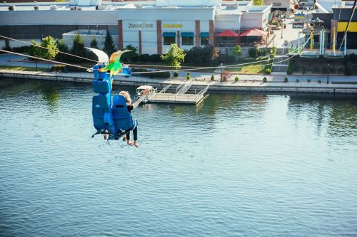 A person rides a blue zip line over a calm body of water, with shops and trees in the background.