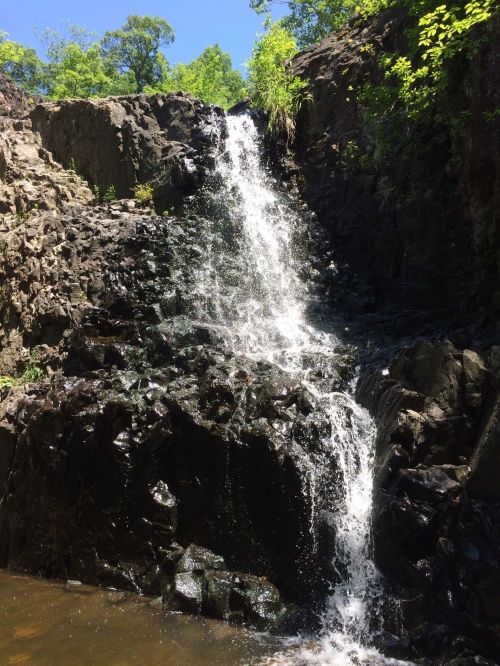 A small waterfall cascading over dark rocks, surrounded by lush green foliage under a clear blue sky.