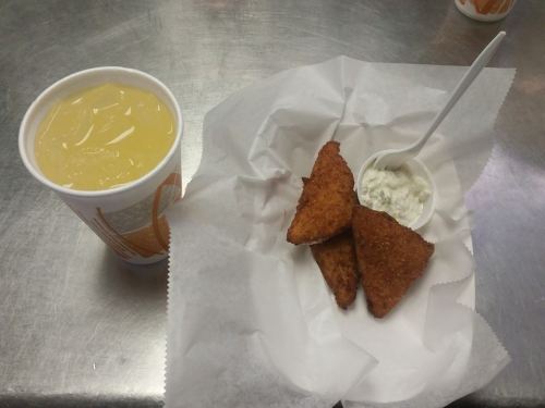 A cup of yellow drink next to a plate of three fried triangle-shaped snacks with a small container of dipping sauce.