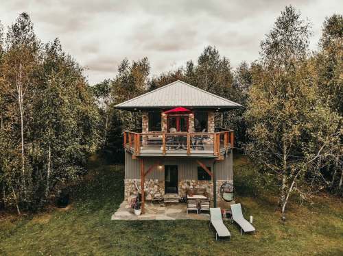 A cozy two-story cabin surrounded by trees, featuring a balcony with a red umbrella and outdoor seating.