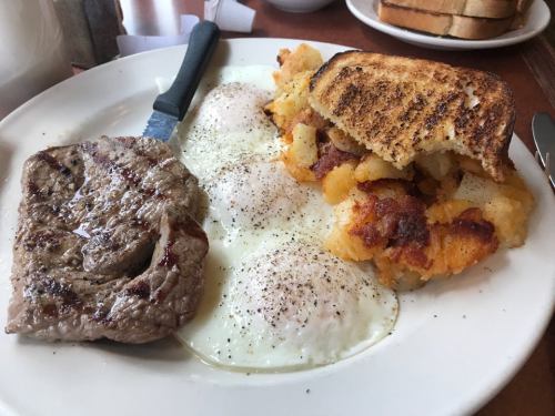 A plate with grilled steak, two sunny-side-up eggs, and crispy hash browns, served with toasted bread.