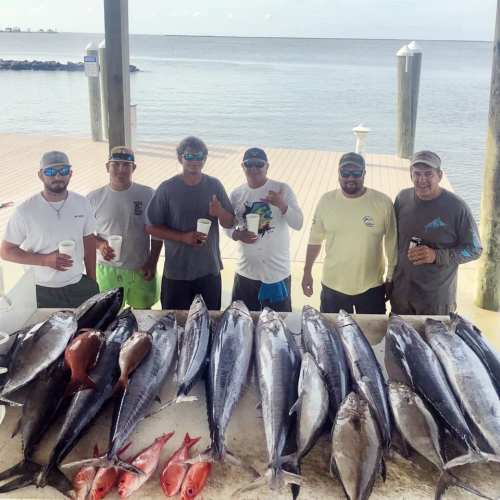 Five fishermen stand by a table displaying a large catch of fish, with a scenic waterfront in the background.