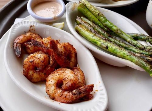 A plate featuring seasoned shrimp and grilled asparagus, accompanied by a small cup of dipping sauce.