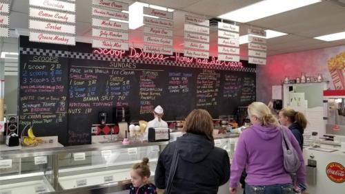 A busy ice cream shop with a menu board, customers at the counter, and a server in a white hat.