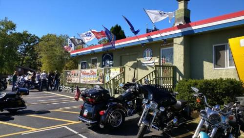 A row of motorcycles parked outside a building with flags and signage, surrounded by greenery and a clear blue sky.
