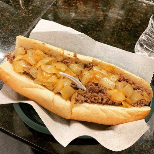 A loaded sandwich with sliced beef, sautéed onions, and a soft hoagie roll, served on a plate with a napkin.