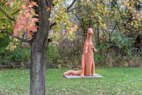A large clay dinosaur sculpture stands in a grassy area surrounded by trees with autumn leaves.