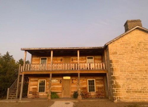A rustic two-story wooden cabin with a stone chimney and a porch, surrounded by trees under a clear sky.