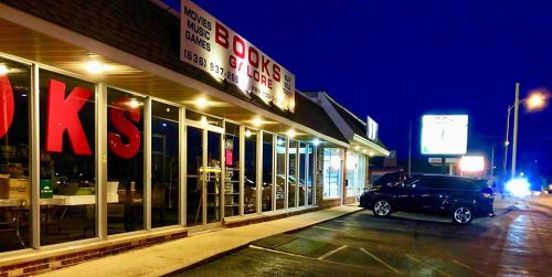 A storefront at night featuring large red letters spelling "BOOKS," with a car parked outside and a lit sign in the background.