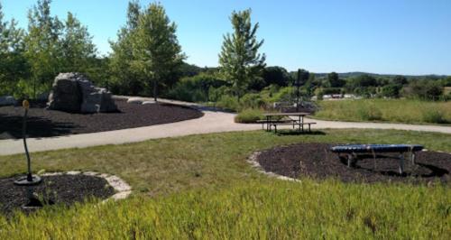 A sunny park scene featuring a walking path, picnic table, and play equipment surrounded by grass and trees.