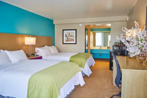 A bright hotel room featuring two beds, a desk, and a colorful accent wall with a vase of flowers.
