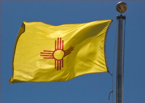 A yellow flag with a red Zia sun symbol, waving against a clear blue sky.