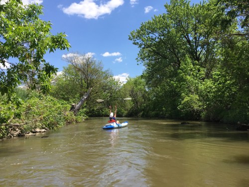 A person paddles a kayak on a calm river surrounded by lush greenery and blue skies with fluffy clouds.