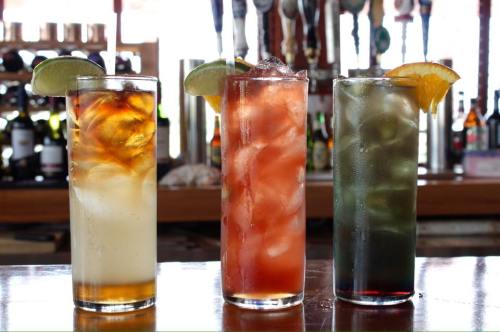 Three tall glasses of colorful cocktails with ice, garnished with lime and orange slices, on a bar counter.