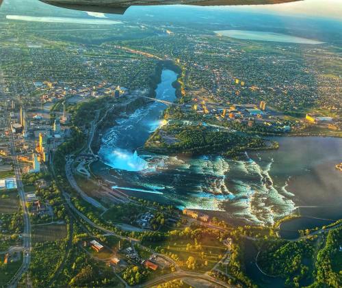 Aerial view of a waterfall surrounded by greenery and a cityscape, with a river flowing into a larger body of water.