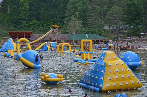 Colorful inflatable water park features slides and climbing structures in a lake, with people swimming and playing.
