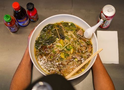 A large bowl of soup with noodles, greens, and meat, surrounded by condiments and a drink on a table.