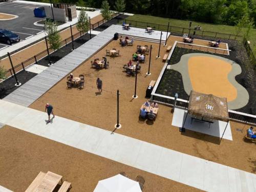 A spacious outdoor area with tables, a playground, and walking paths, surrounded by greenery and a parking lot.