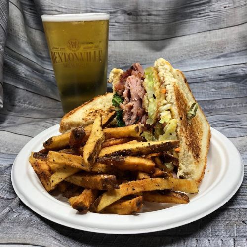 A hearty sandwich with layers of meat and veggies, served with crispy fries and a glass of beer in the background.