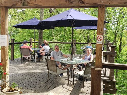 A wooden deck with tables and umbrellas, where people are dining among trees in a lush green setting.