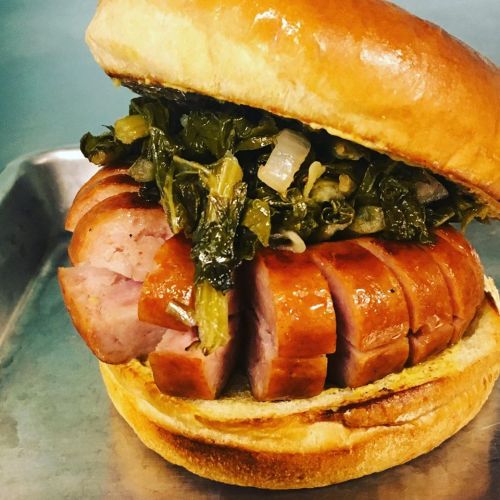 A close-up of a sandwich featuring sliced sausage topped with greens on a soft bun, served on a metal tray.