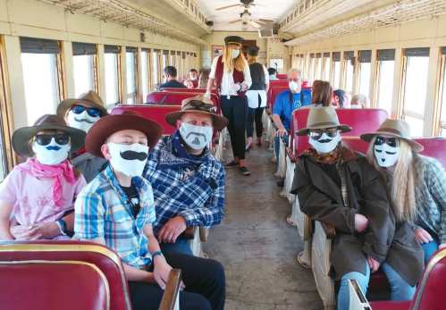 A group of people wearing hats and masks with mustache designs sit inside a vintage train car, smiling at the camera.