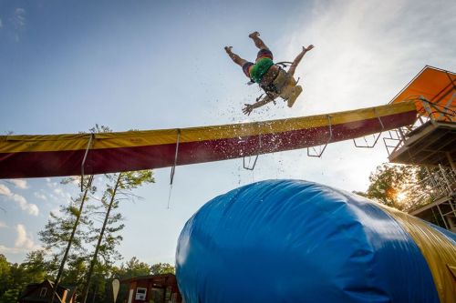 A person jumps off a water slide, splashing into a large inflatable landing pad below, with trees and a blue sky in the background.