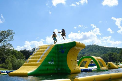 Two children jump off a large inflatable water slide into a lake on a sunny day, surrounded by greenery.