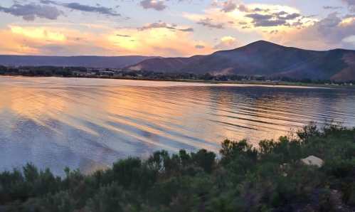 Serene lake at sunset, reflecting colorful clouds and mountains in the background, with gentle ripples on the water.
