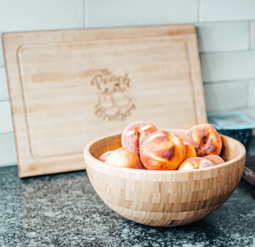 A wooden bowl filled with peaches sits on a countertop next to a engraved wooden lid that reads "The Peach Brokerage."