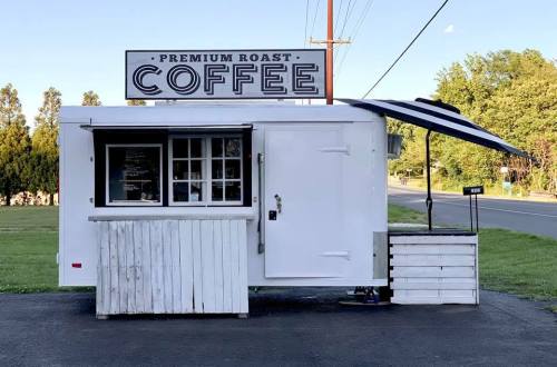 A white coffee stand with a sign reading "Premium Roast Coffee," featuring an awning and a grassy area nearby.