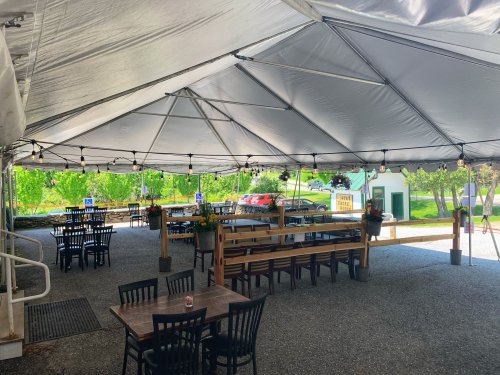 A spacious outdoor dining area under a large tent, featuring tables, chairs, and greenery in the background.