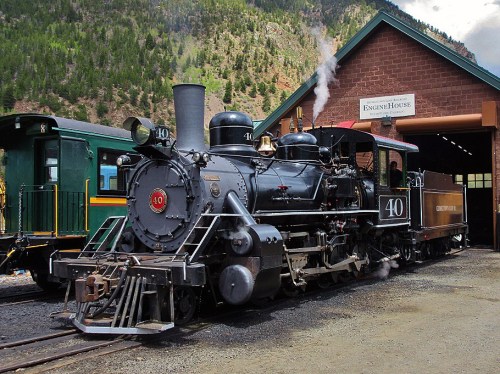 A vintage steam locomotive numbered 40, with smoke billowing, parked outside a brick engine house in a mountainous area.
