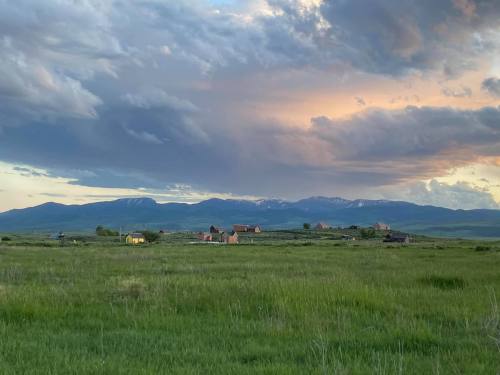 A serene landscape featuring a grassy field, distant mountains, and a small village under a cloudy sky at sunset.