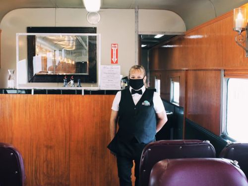 A masked server stands in a vintage train car, featuring wooden paneling and elegant decor.