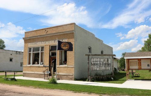 Historic building of Home State Bank with a sign for Sandhills Heritage, surrounded by green grass and blue sky.