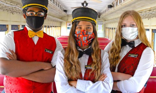 Three train conductors in masks stand confidently inside a vintage train car, wearing red vests and black hats.