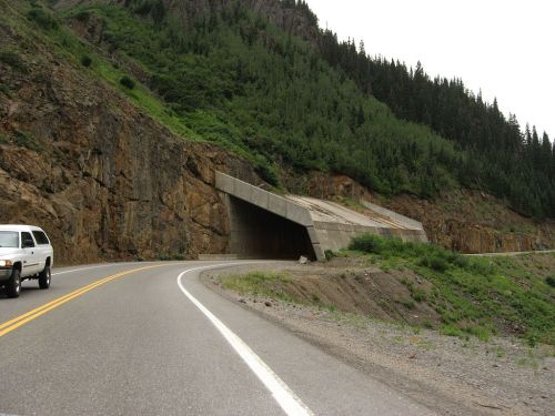 A concrete wildlife overpass on a winding road, surrounded by rocky terrain and dense green forest.