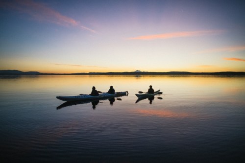 Three kayakers paddle on calm water at sunset, with a mountain silhouette in the background and colorful sky reflections.